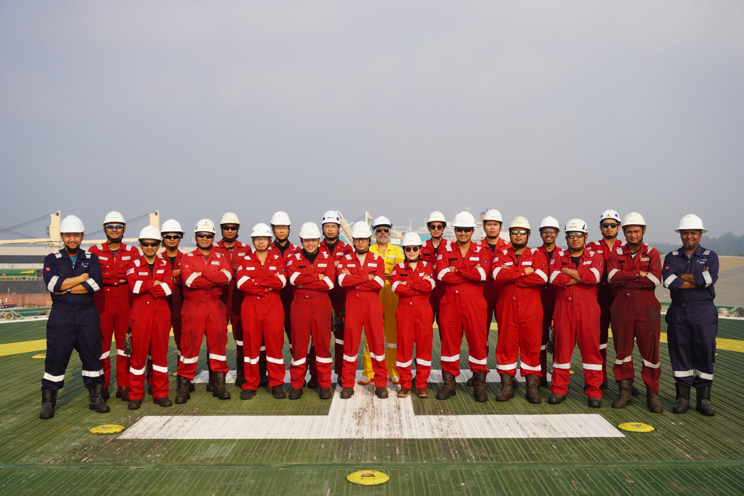 Group of workers in red coveralls and white helmets posing on a ship or platform deck outdoors.